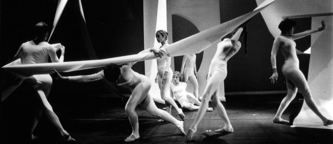 Seven dancers holding white paper rolls