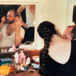 Thumbnail of woman with braid applying makeup to a man seated